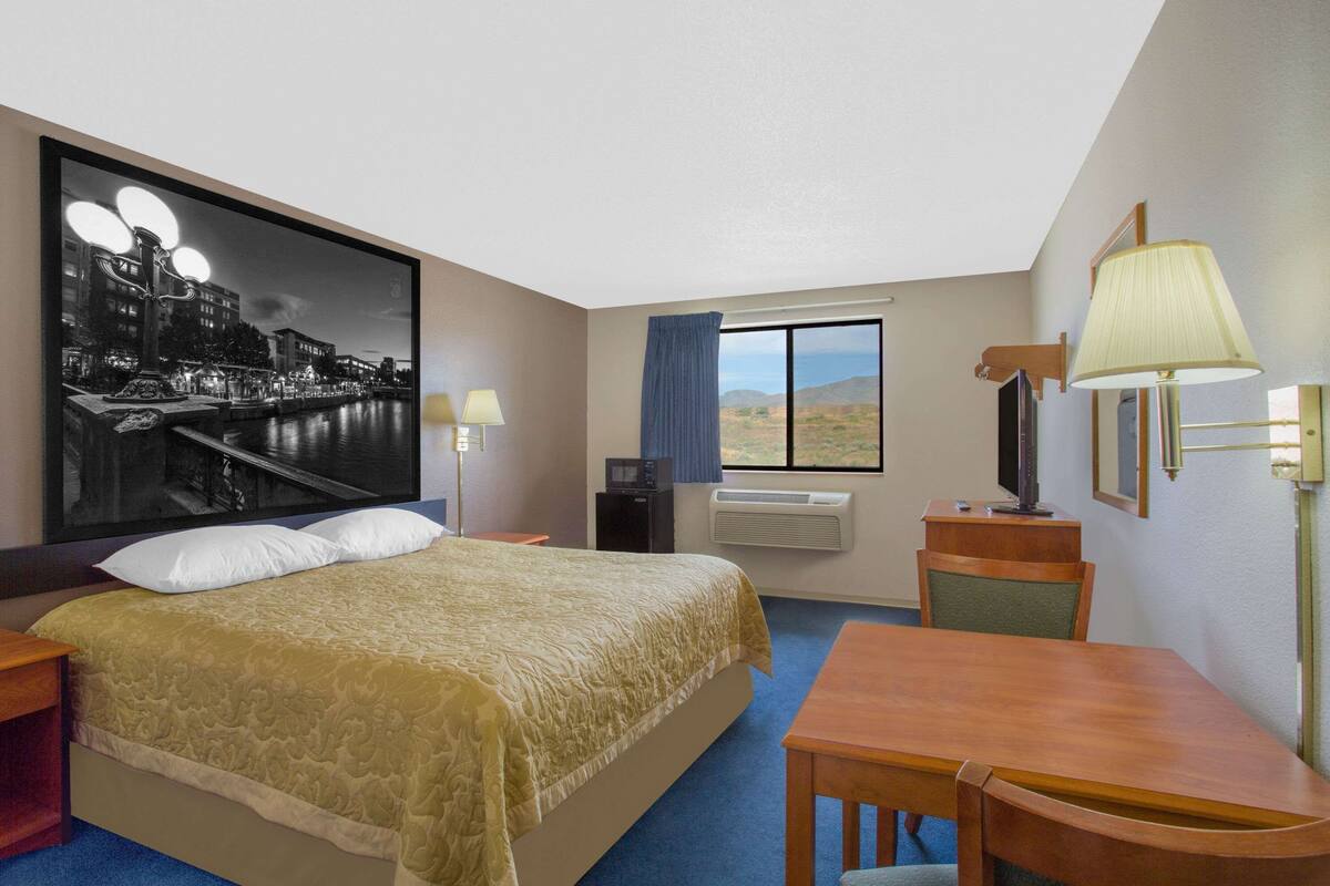 Une chambre d’hôtel avec un grand lit, un bureau, une chaise, une télévision, une fenêtre donnant sur le désert et un tableau représentant un paysage urbain.