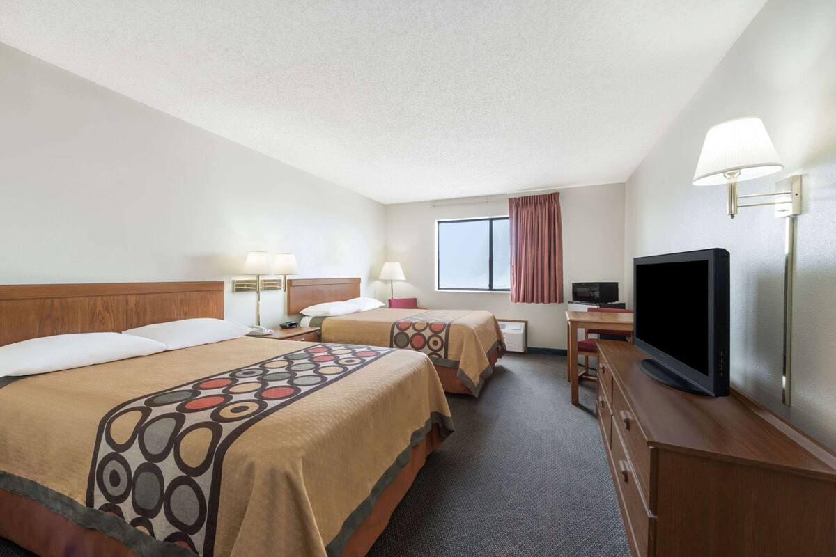 Ein Hotelzimmer mit zwei Betten, einem Schreibtisch, einem Fernseher und einem fensterbelaubten Fenster.