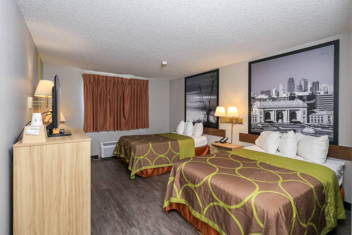 Ein Hotelzimmer mit zwei Betten, einem Schreibtisch, einer Lampe, einem großen Stadtplakat und einem Fenster mit braunen Vorhängen.