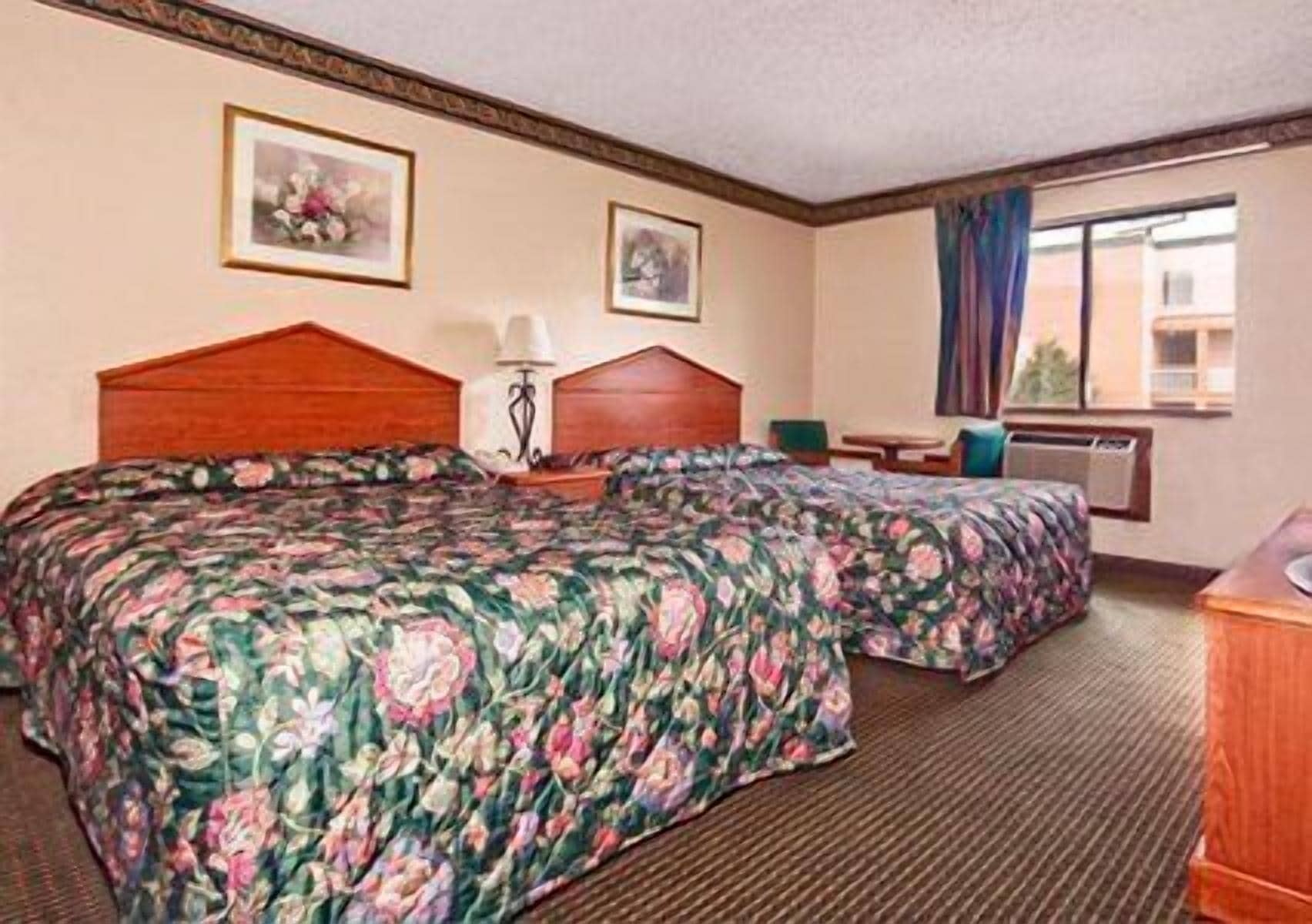 A hotel room with two beds, a desk, a chair, a lamp, and a window with curtains.