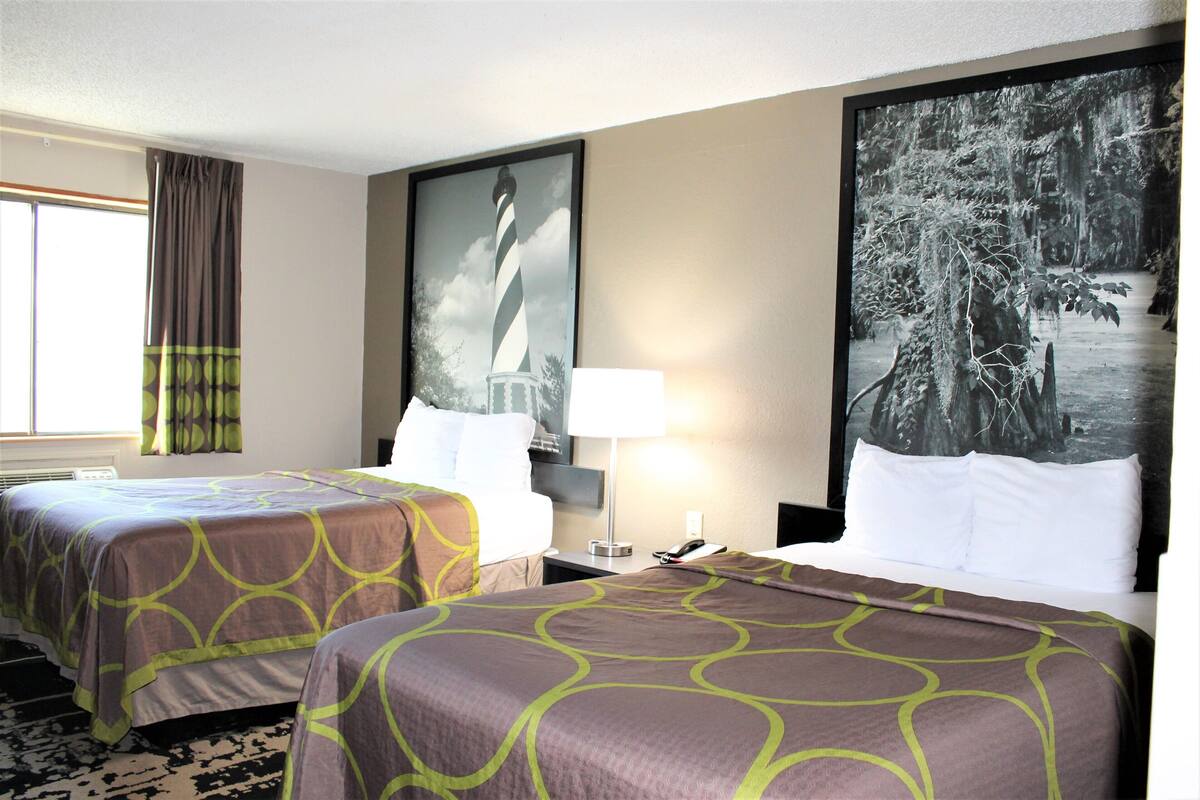 Une chambre d’hôtel avec deux lits, un grand tableau encadré représentant un phare, une lampe et une fenêtre avec des rideaux.