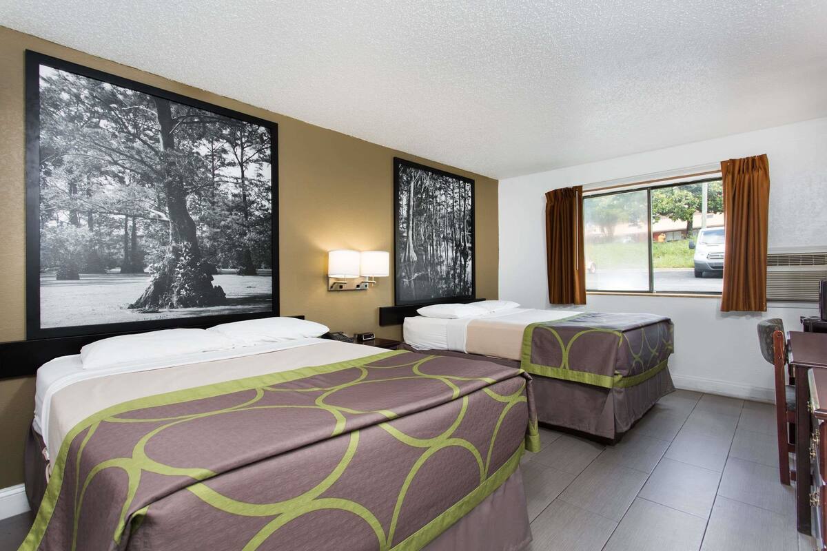 Une chambre d’hôtel avec deux lits, un bureau, une chaise, une fenêtre avec des rideaux et deux grandes images accrochées au mur.