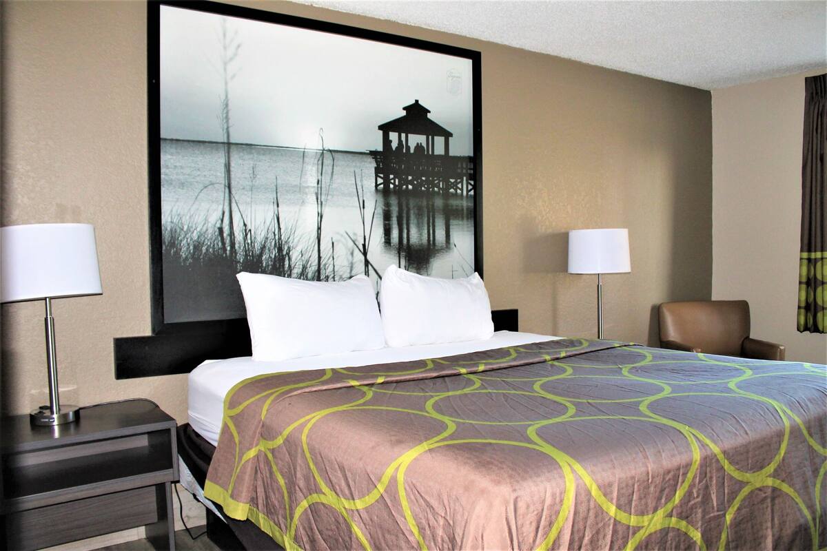 Une chambre d’hôtel avec un lit, deux lampes de chevet, une chaise et un grand tableau accroché au mur représentant une jetée au-dessus de l’eau.