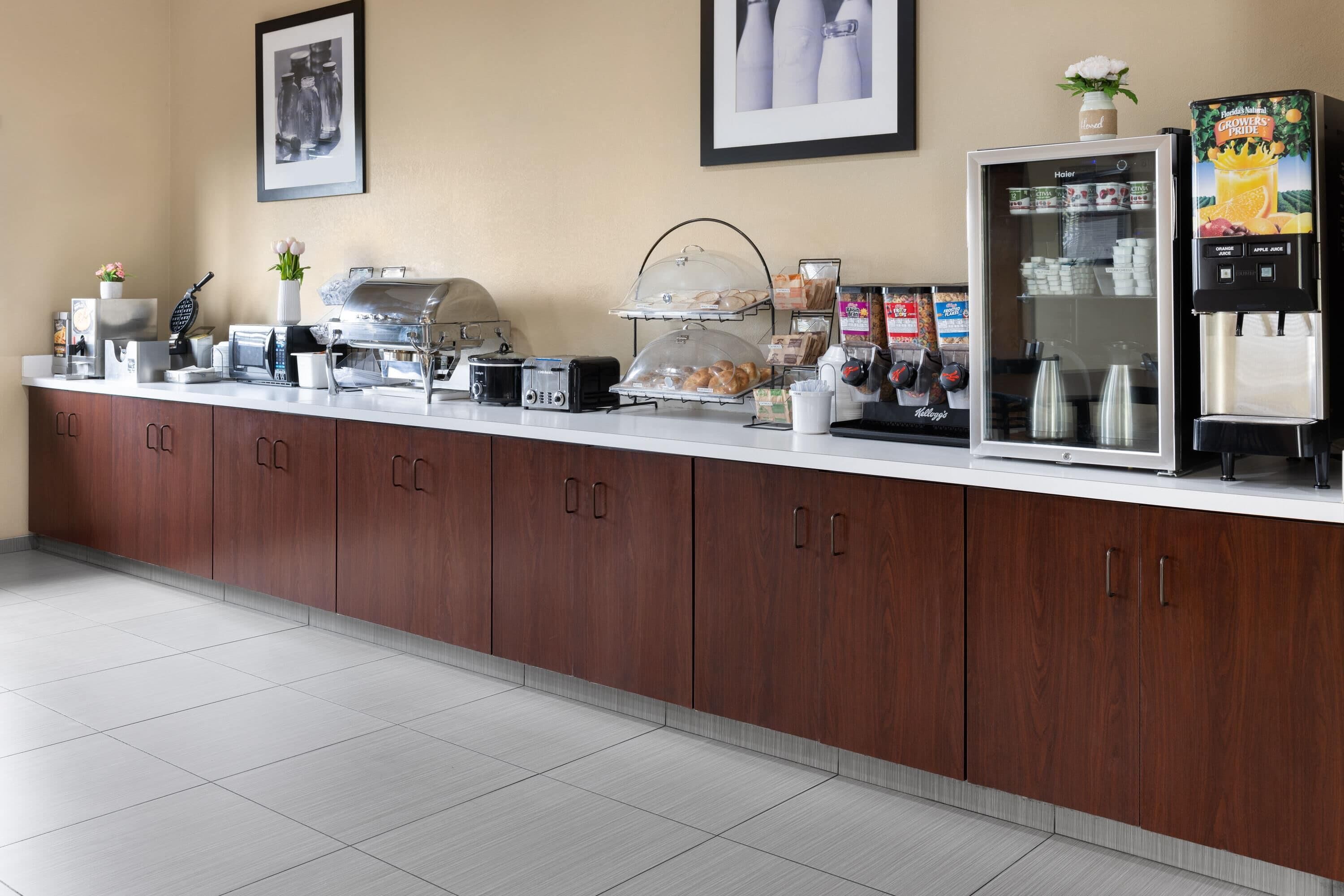 A hotel breakfast area with a coffee machine, toaster, kettle, and a refrigerator with a beverage dispenser.