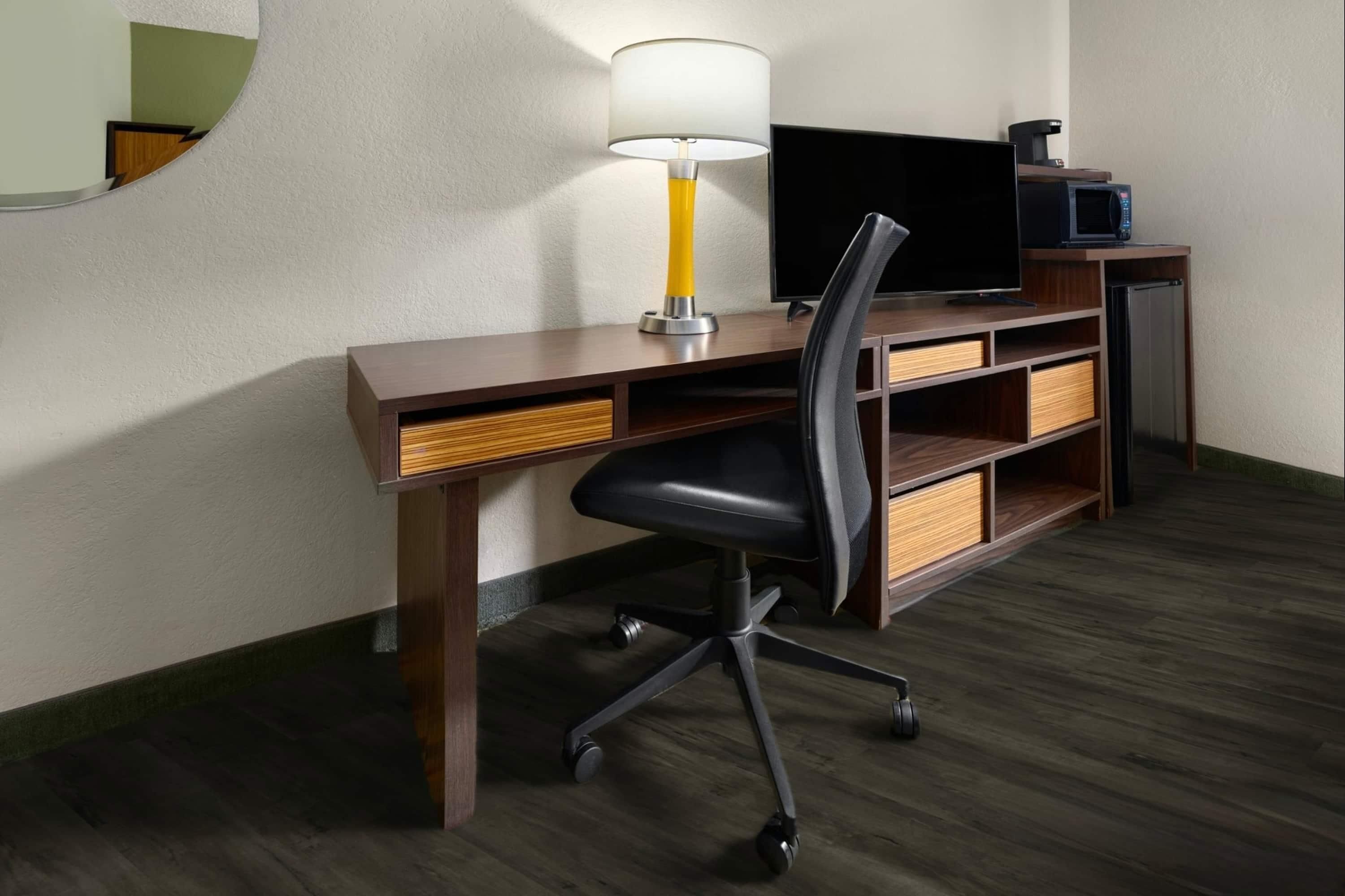 Ein Hotelzimmer mit einem Holztisch, einem schwarzen Bürostuhl, einem Fernseher, einer Mikrowelle und einer Kaffeemaschine.