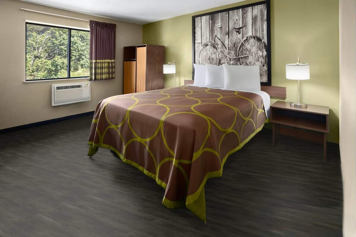 A hotel room with a large bed, a window with curtains, a wall-mounted air conditioner, a nightstand with a lamp, and a painting on the wall.