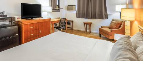 A hotel room with a bed, a desk with a chair, a television, a dresser, a small table, a lamp, and a window with curtains.