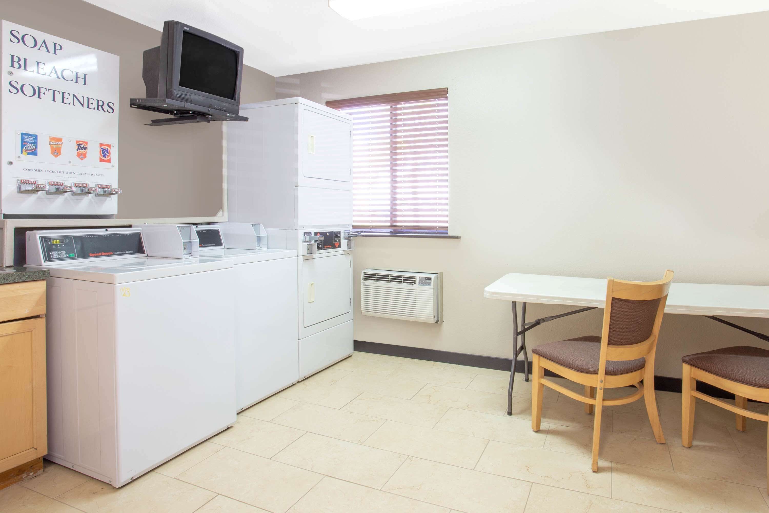 Une buanderie équipée d’un lave-linge, d’un sèche-linge et d’une planche à repasser. Un téléviseur est fixé au mur et il y a une fenêtre avec des stores. Deux chaises en bois sont placées près d’une table.
