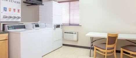 Una sala de lavandería con lavadora, secadora y plancha. Hay un televisor montado en la pared y una ventana con persianas. Dos sillas de madera están colocadas cerca de una mesa.