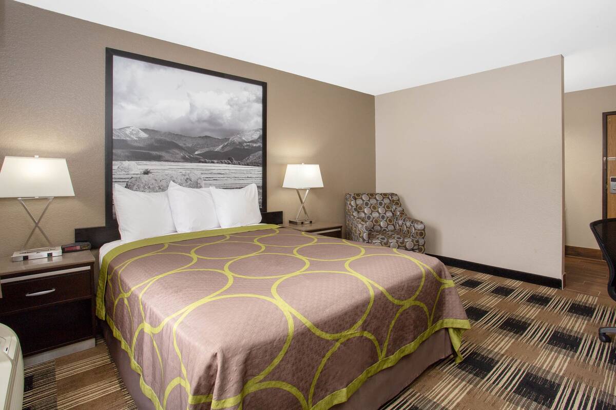 Een hotelkamer met een groot bed, twee nachtkastjes met lampen, een stoel en een groot, ingelijst schilderij aan de muur.
