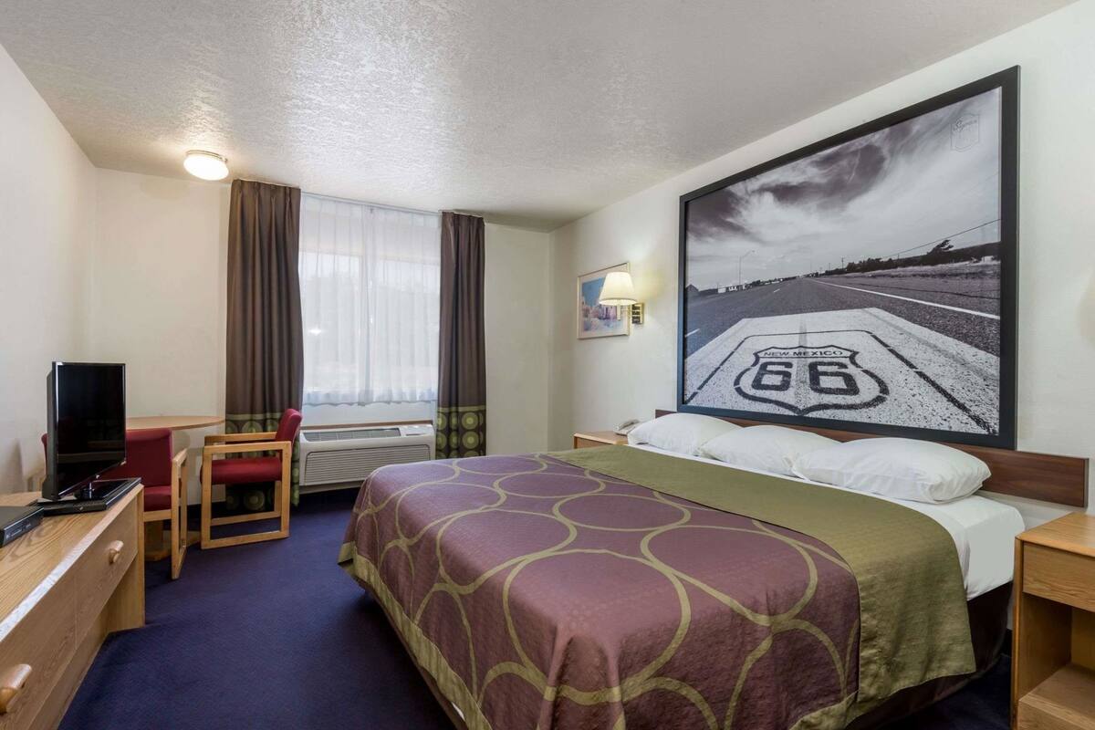 A hotel room with a bed, a desk with a chair, a television, a window with curtains, and a large painting of a road.