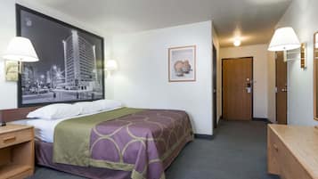 A hotel room with a bed, a nightstand, a wooden dresser, a framed picture on the wall, and a door leading to another room.