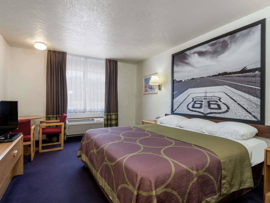 A hotel room with a bed, a desk with a computer, a chair, a window with curtains, and a wall-mounted TV.