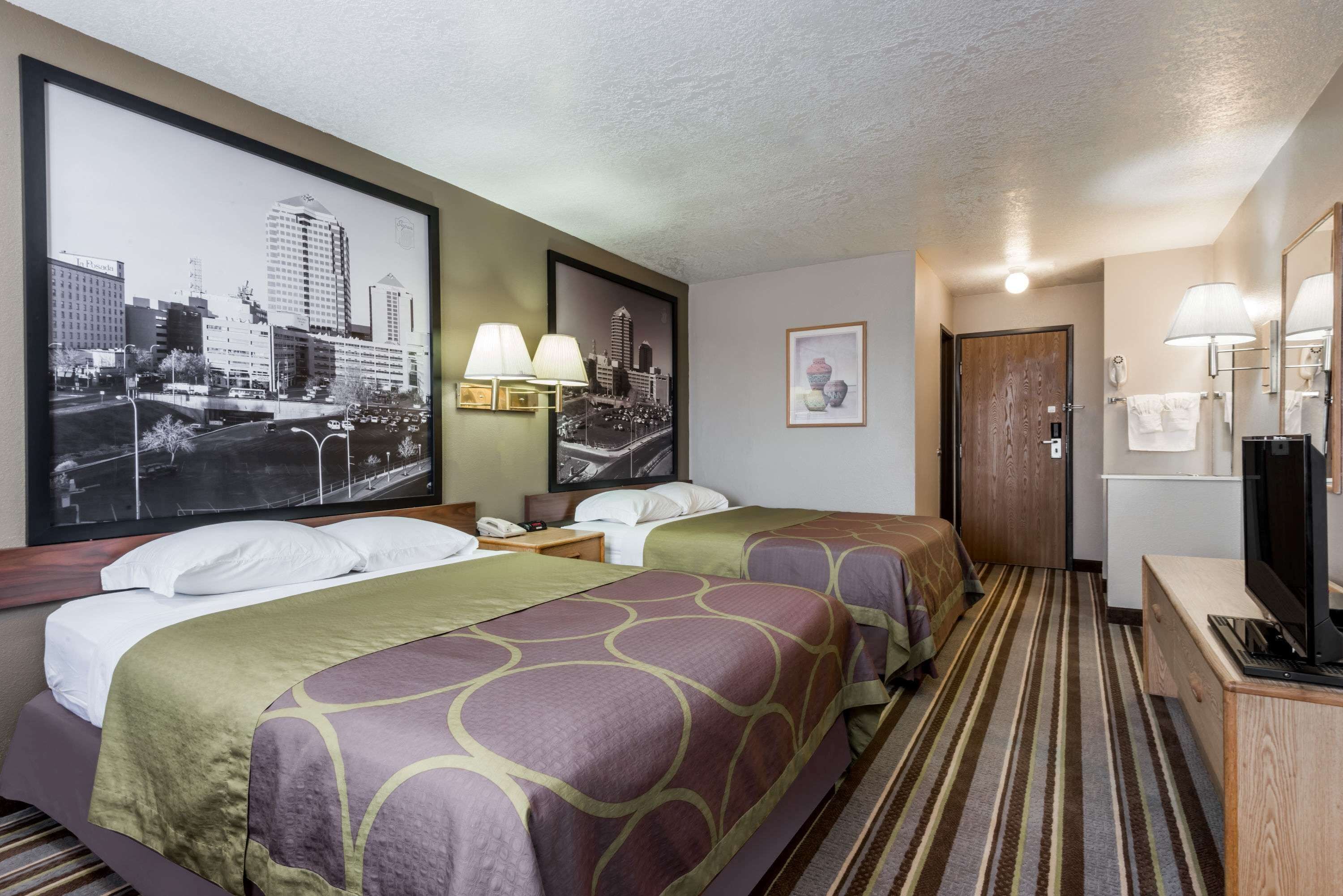 A hotel room with two beds, a cityscape poster, a framed picture, a desk with a TV, and a bathroom door.
