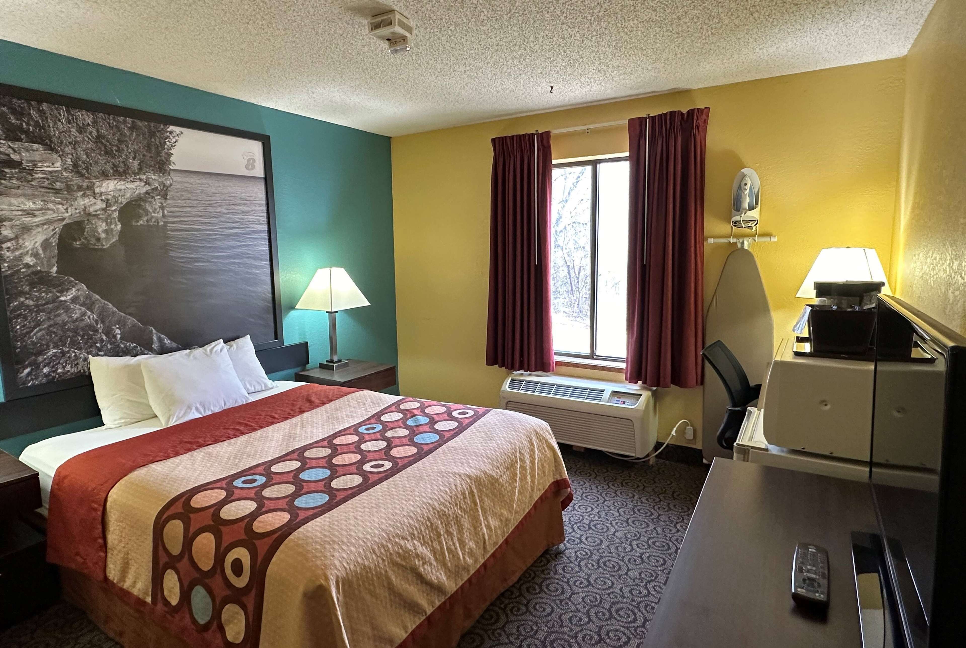 A hotel room with a bed, a desk, an ironing board, a lamp, and a window with curtains.