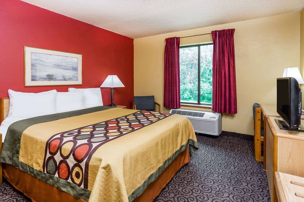 Habitación de hotel con cama, escritorio con televisor, silla, ventana con cortinas y unidad de aire acondicionado.