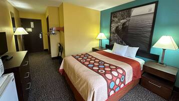 A hotel room with a bed, two bedside tables with lamps, a desk with a lamp, a refrigerator, a door, and a wall with a painting.
