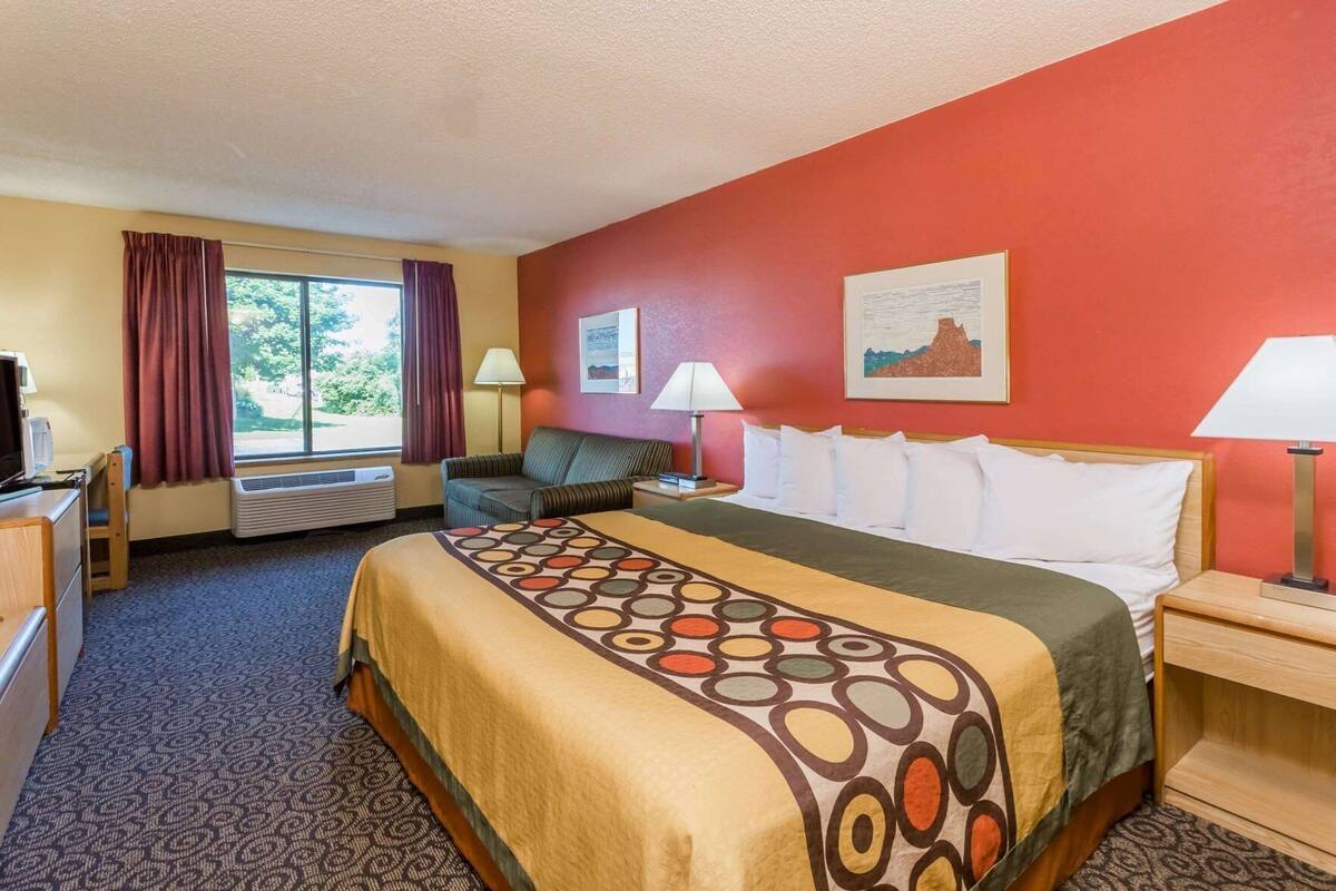 Una habitación de hotel con cama, sofá, escritorio, ventana con cortinas y una pared con dos cuadros enmarcados.