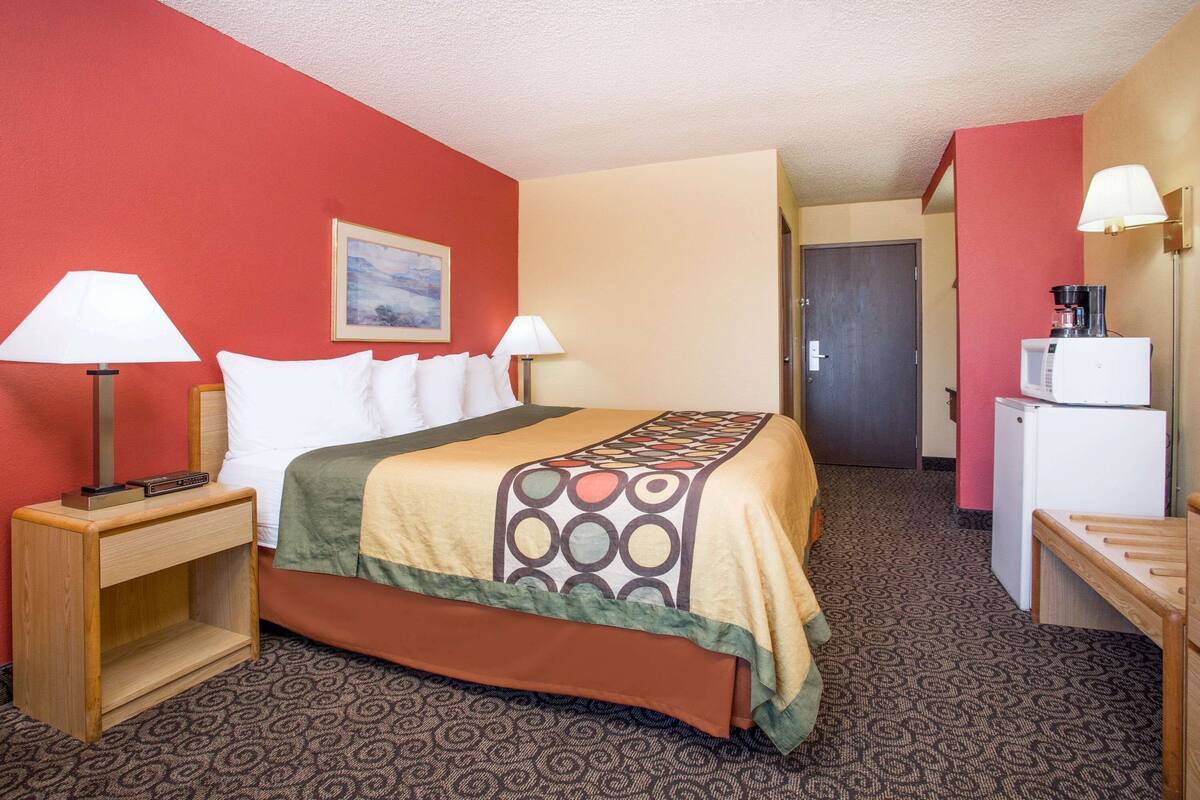 Habitación de hotel con una cama, dos lámparas de noche, una mesita con lámpara, un microondas y un refrigerador.