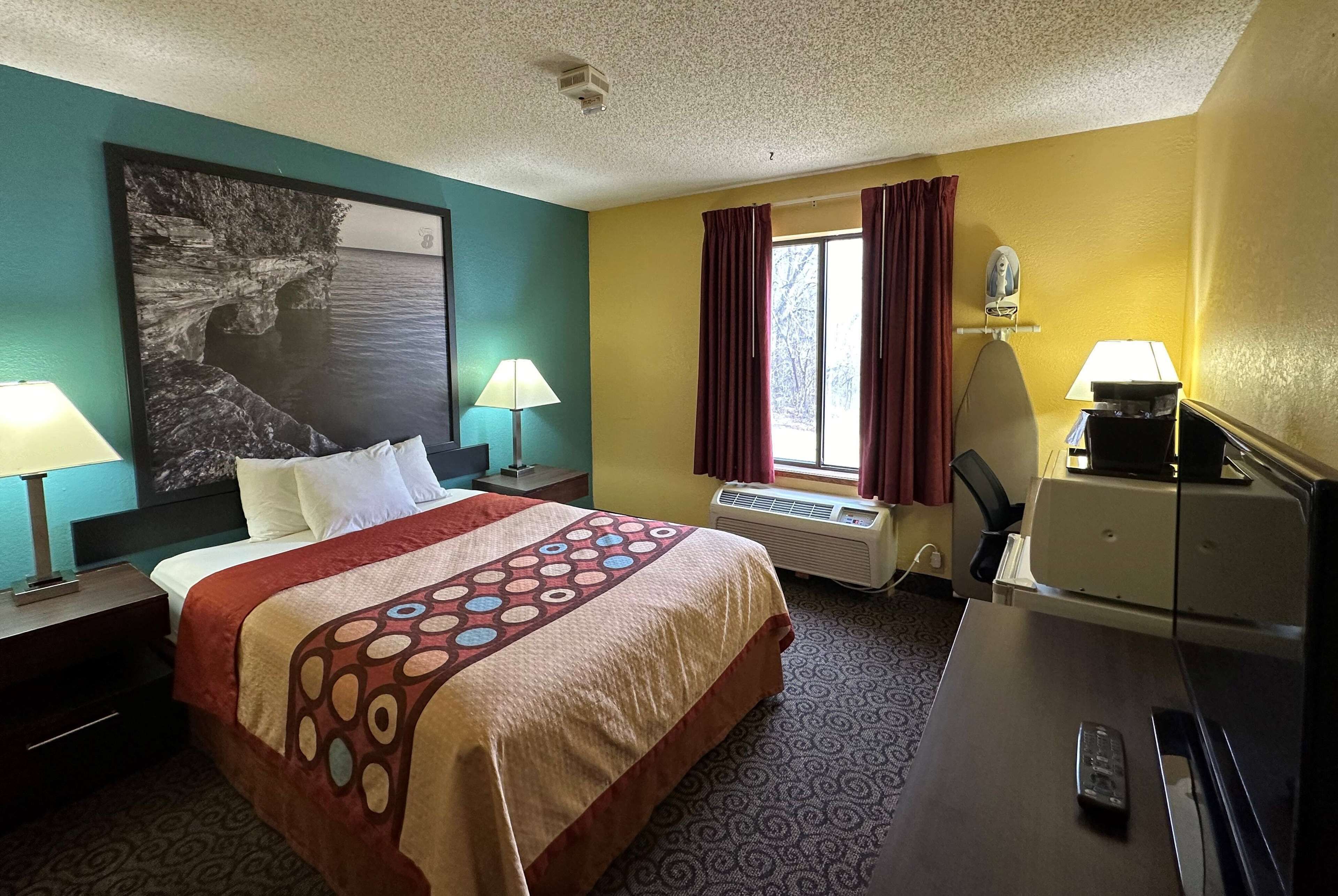 A hotel room with a bed, two bedside lamps, a desk with a computer, a window with curtains, and a large painting on the wall.