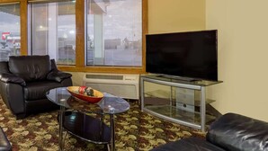 A living room with a leather sofa, a glass coffee table, a TV on a glass cabinet, and a window with blinds.