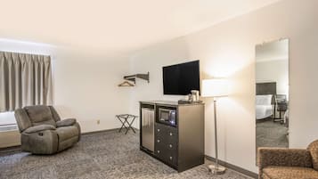 A hotel room with a gray recliner, a minibar, a flat-screen TV, a standing lamp, and a large mirror.