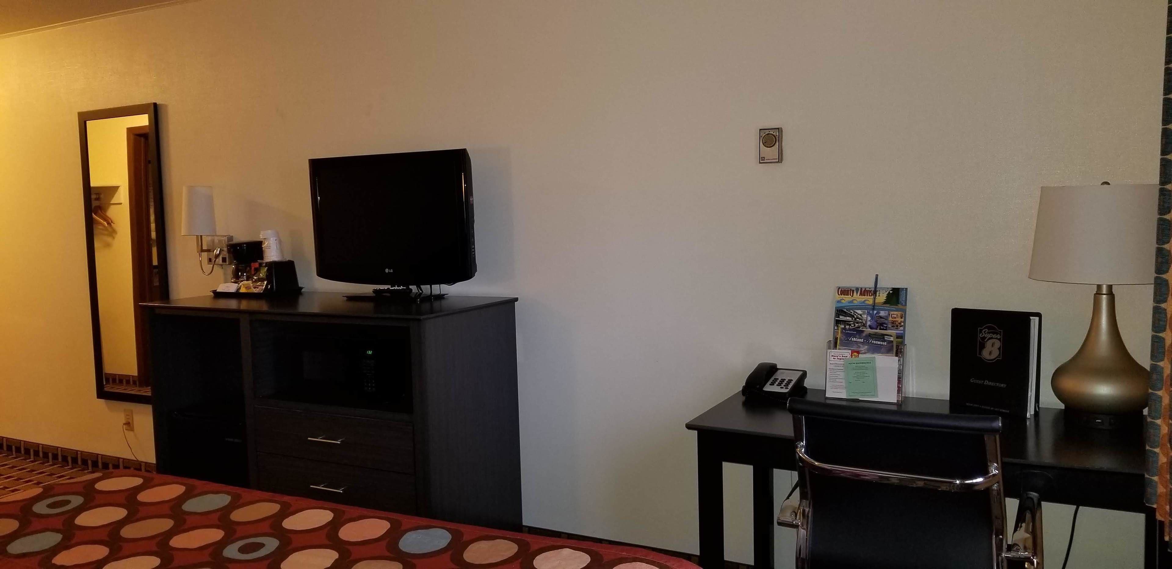 A hotel room with a bed, a television on a stand, a desk with a lamp, a telephone, and a small table with brochures.