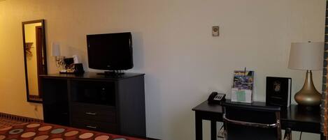 A hotel room with a bed, a television on a stand, a desk with a lamp, a telephone, and a small table with brochures.