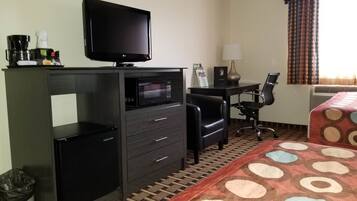 A hotel room with a flat-screen TV, a microwave, a desk with a lamp, a chair, and a patterned bedspread.