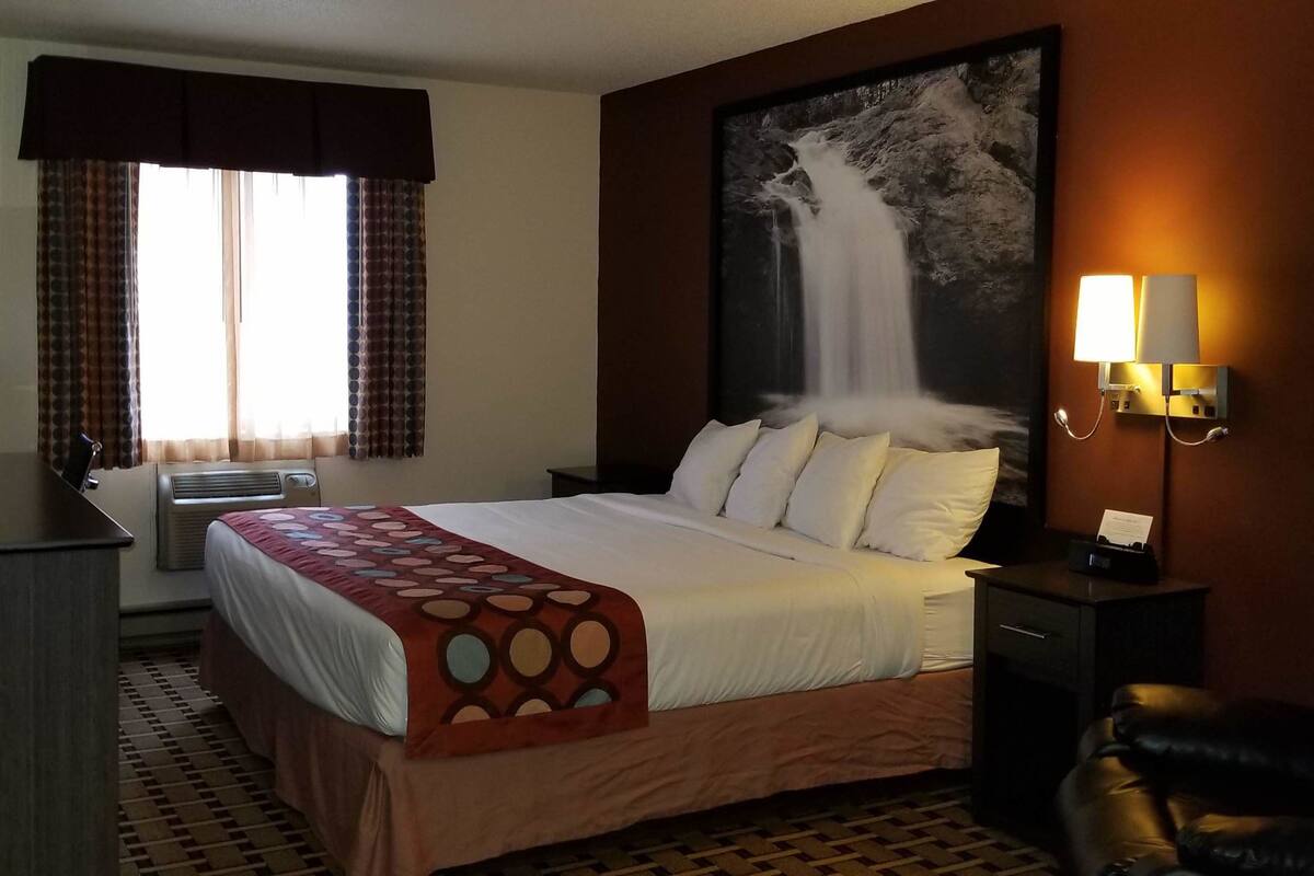Une chambre d’hôtel comprenant un lit, un bureau avec une lampe, un téléviseur fixé au mur, un grand tableau représentant une cascade et une table de chevet avec une lampe.