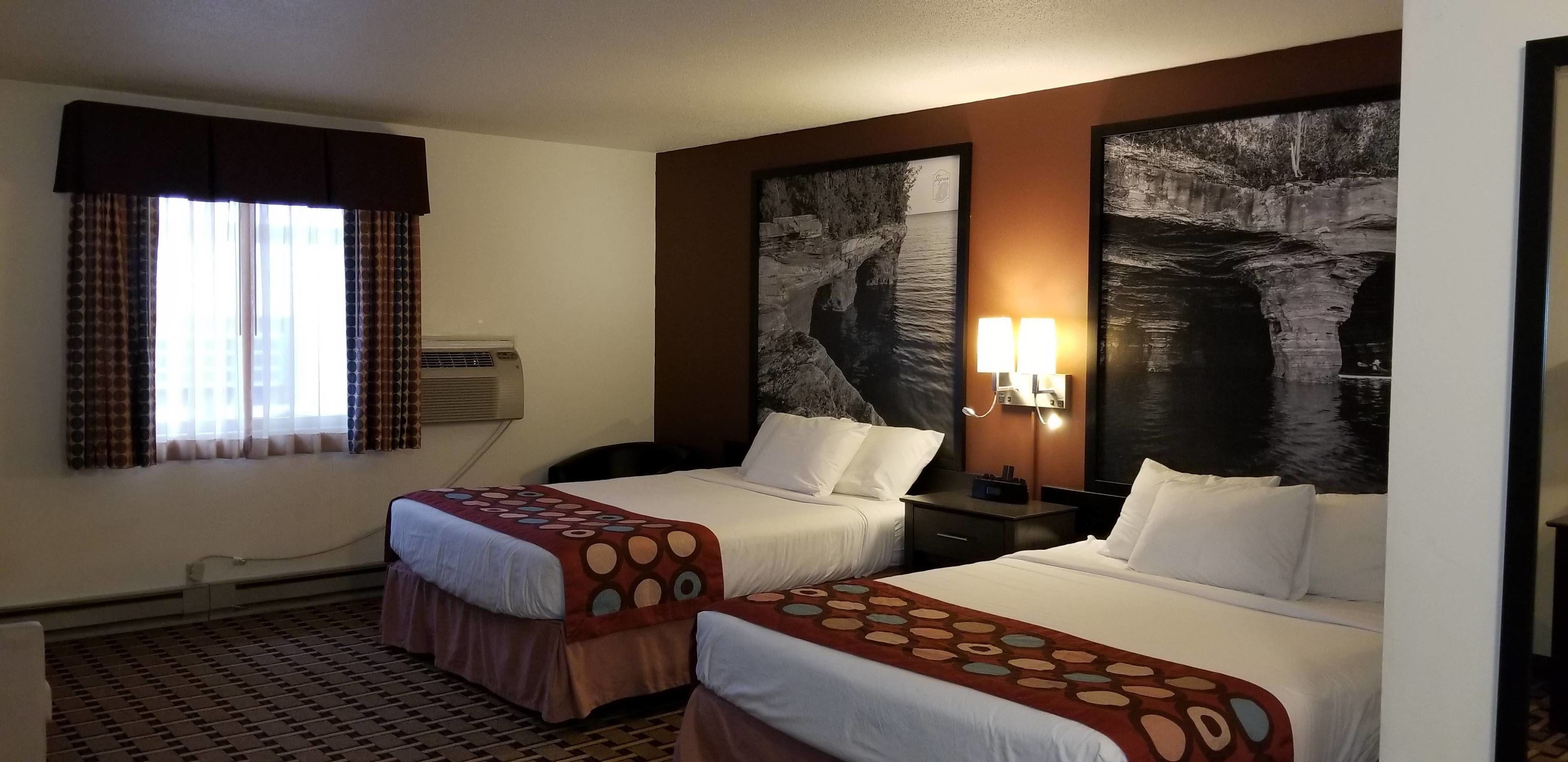 A hotel room with two beds, a window with curtains, an air conditioning unit, and a wall-mounted lamp.