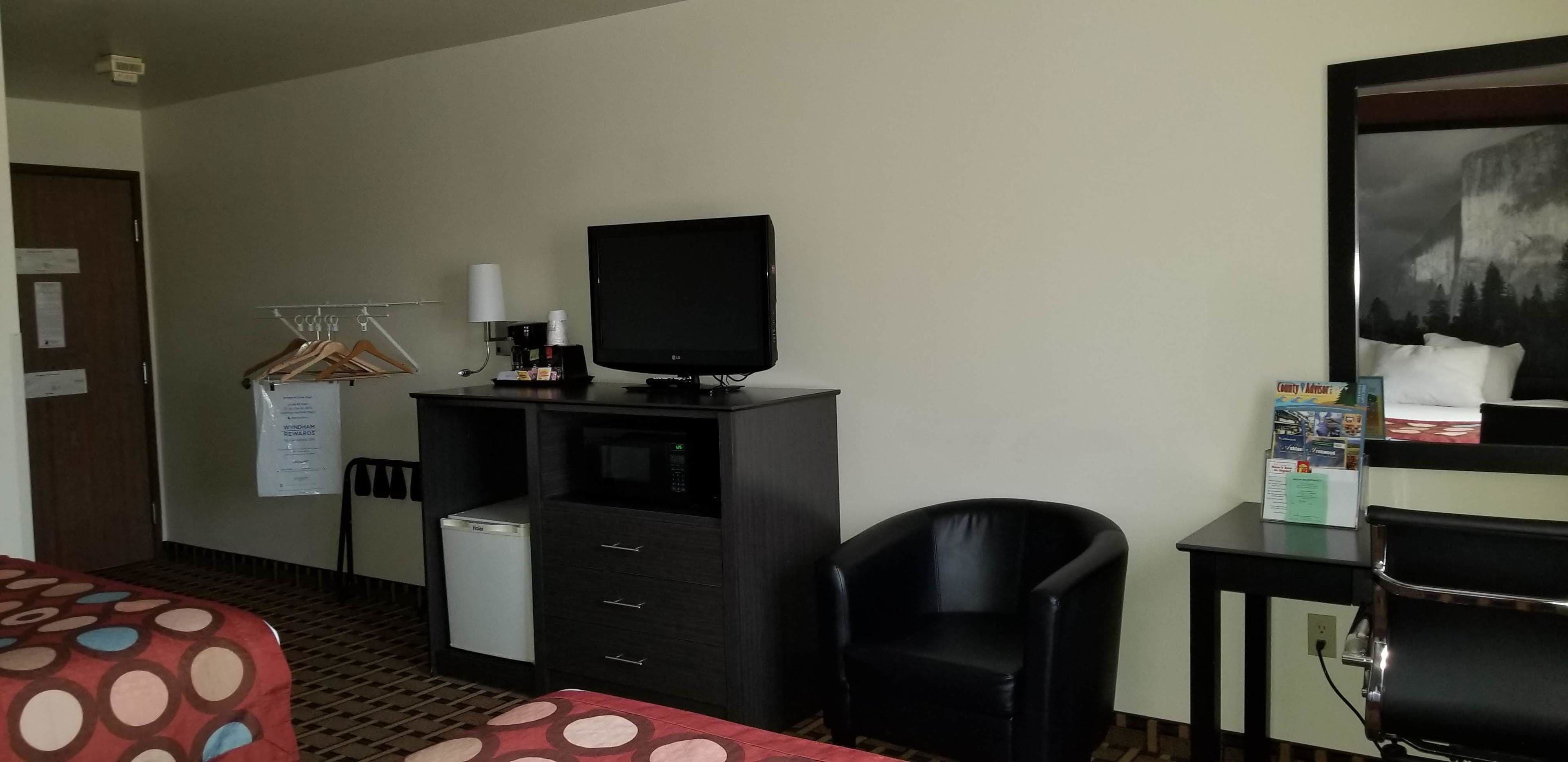 A hotel room with a bed, a desk with a television, a refrigerator, a chair, and a small table with a pillow and a magazine.