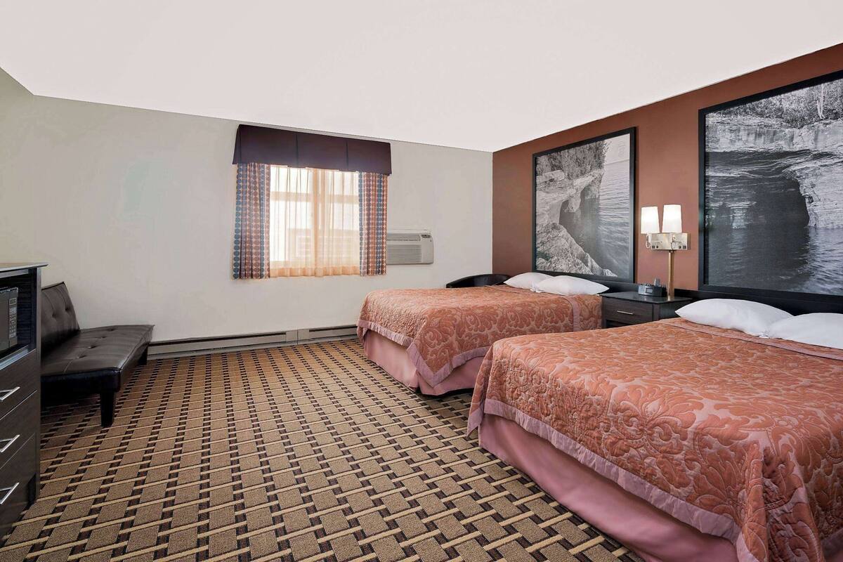 Une chambre d’hôtel avec deux lits, un bureau, une chaise, une fenêtre avec des rideaux et des œuvres d’art accrochées aux murs.