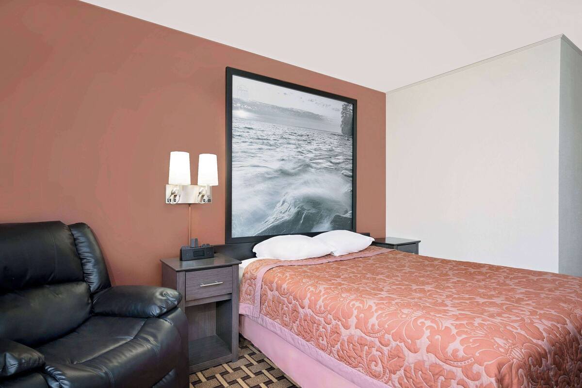 Une chambre d’hôtel avec un lit, un fauteuil en cuir noir, une table de chevet avec deux lampes et un tableau encadré au mur.
