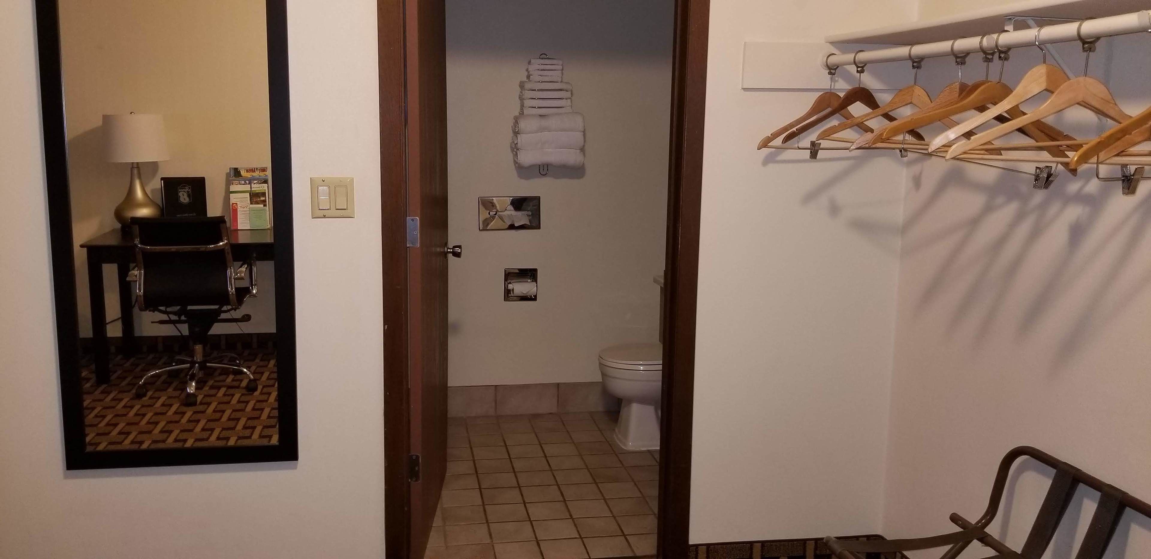 Una camera d'albergo con una sedia in legno, una scrivania con una lampada, uno specchio, un bagno con il water, un portaservizi e un appendiabiti.