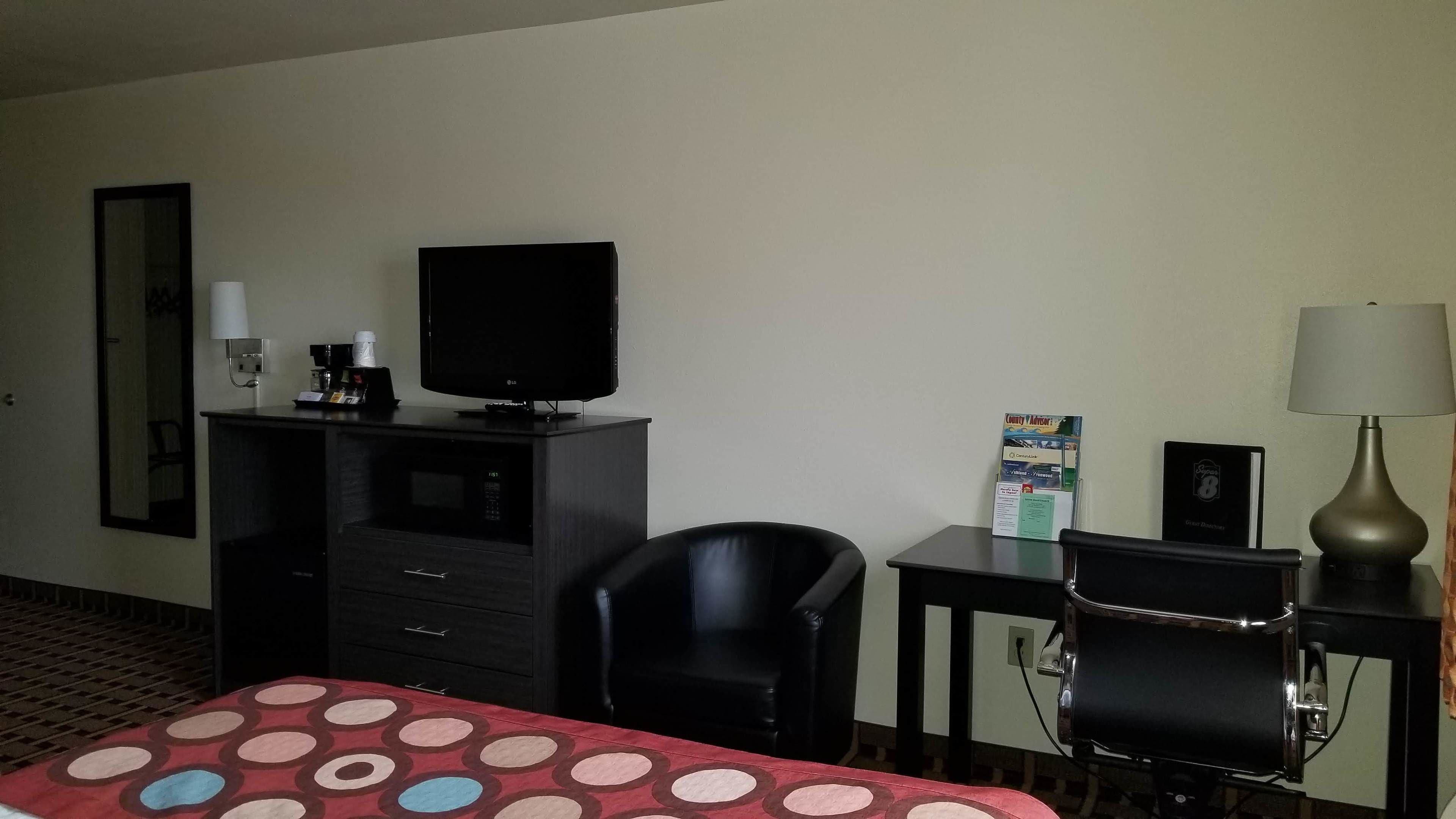 A hotel room with a bed, a desk with a television, a chair, a lamp, and a small table with a brochure.