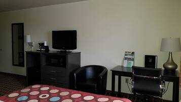 A hotel room with a bed, a desk with a television, a chair, a lamp, and a small table with a brochure.