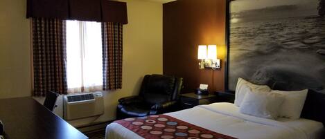 A hotel room with a bed, a desk, a chair, an air conditioner, a wall-mounted lamp, and a painting of a seascape.