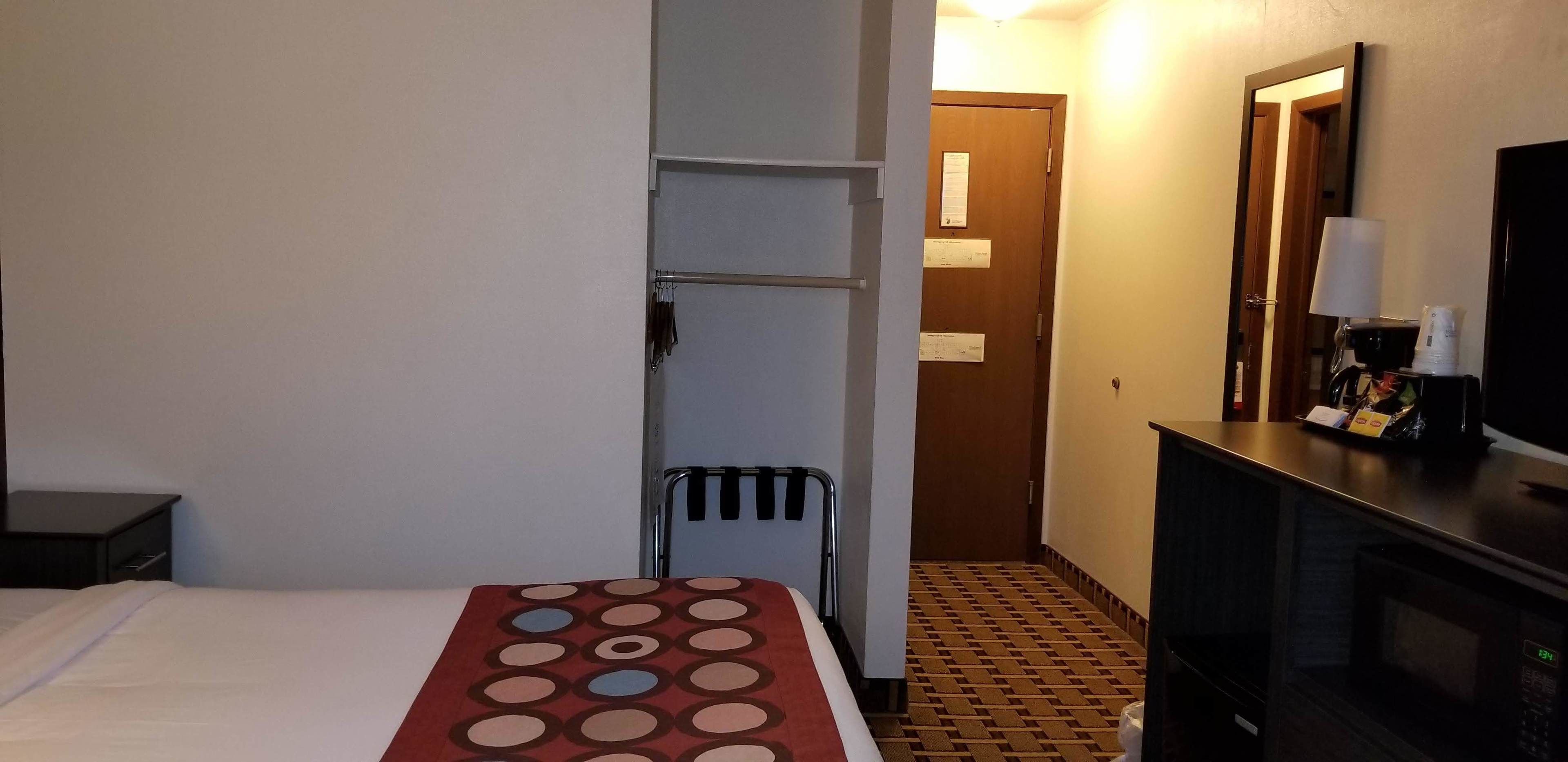 A hotel room with a bed, a small table with a patterned tablecloth, a chair, a wardrobe, a microwave, and a television on a cabinet.