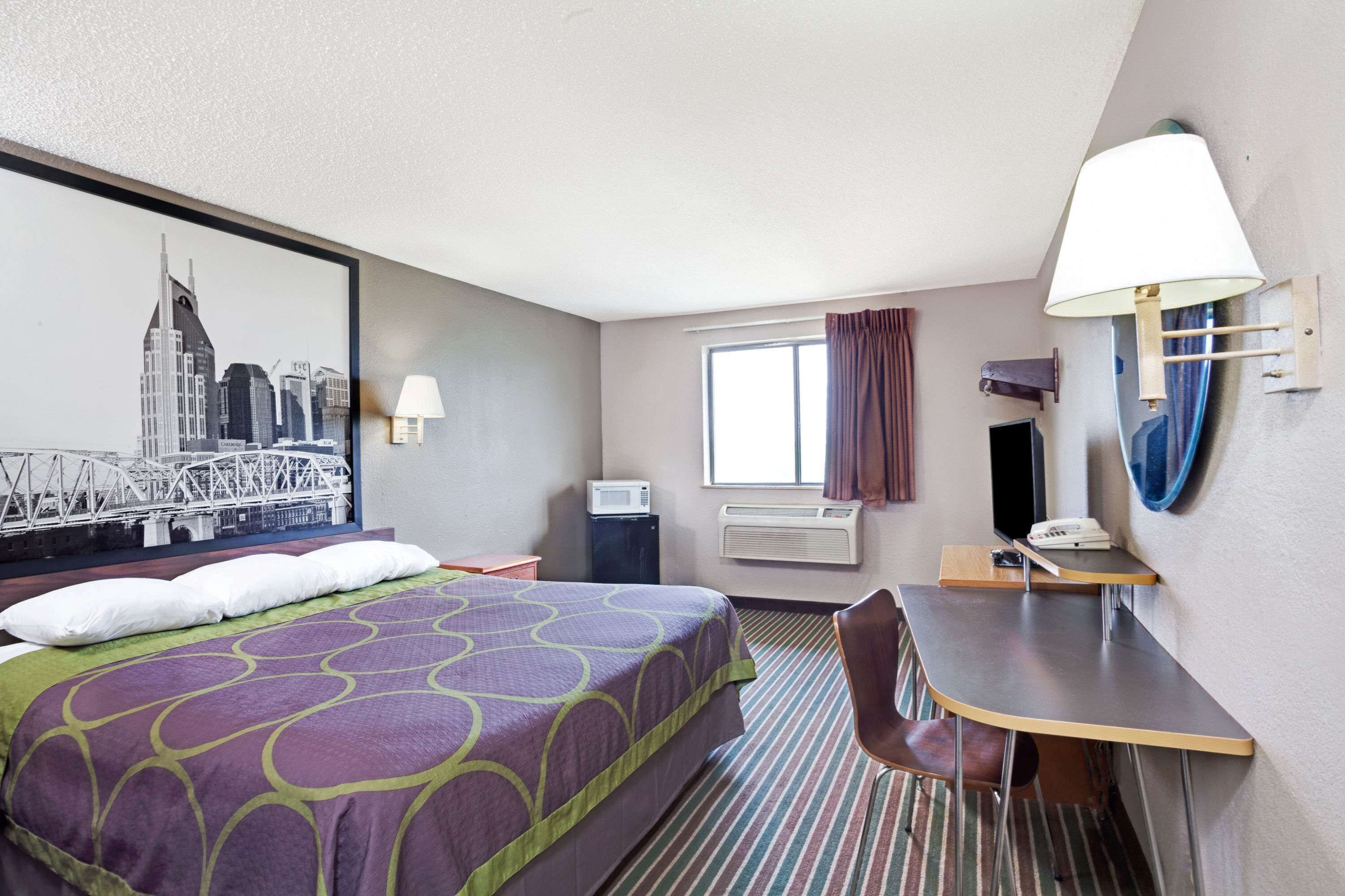 A hotel room with a bed, a desk, a chair, a window with curtains, a wall-mounted TV, and a cityscape painting.
