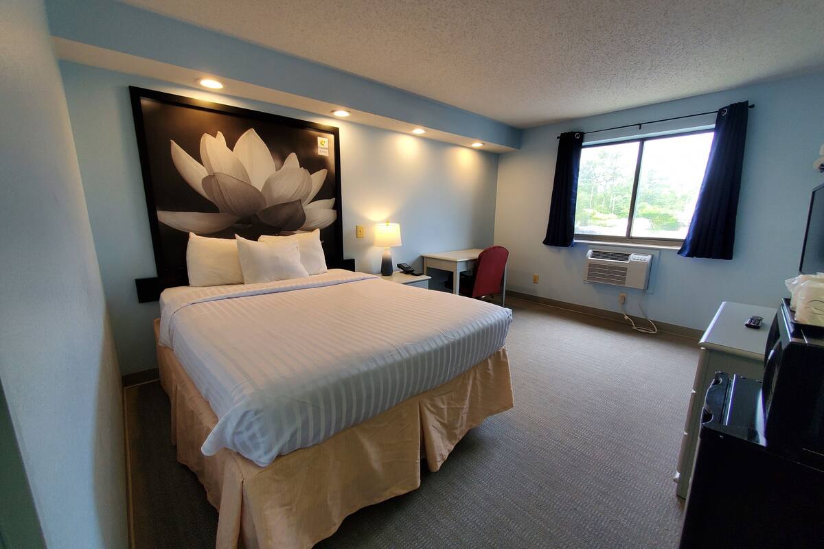Ein Hotelzimmer mit einem großen Bett, einem Schreibtisch, einem Stuhl, einer Lampe und einem Vorhangfenster.