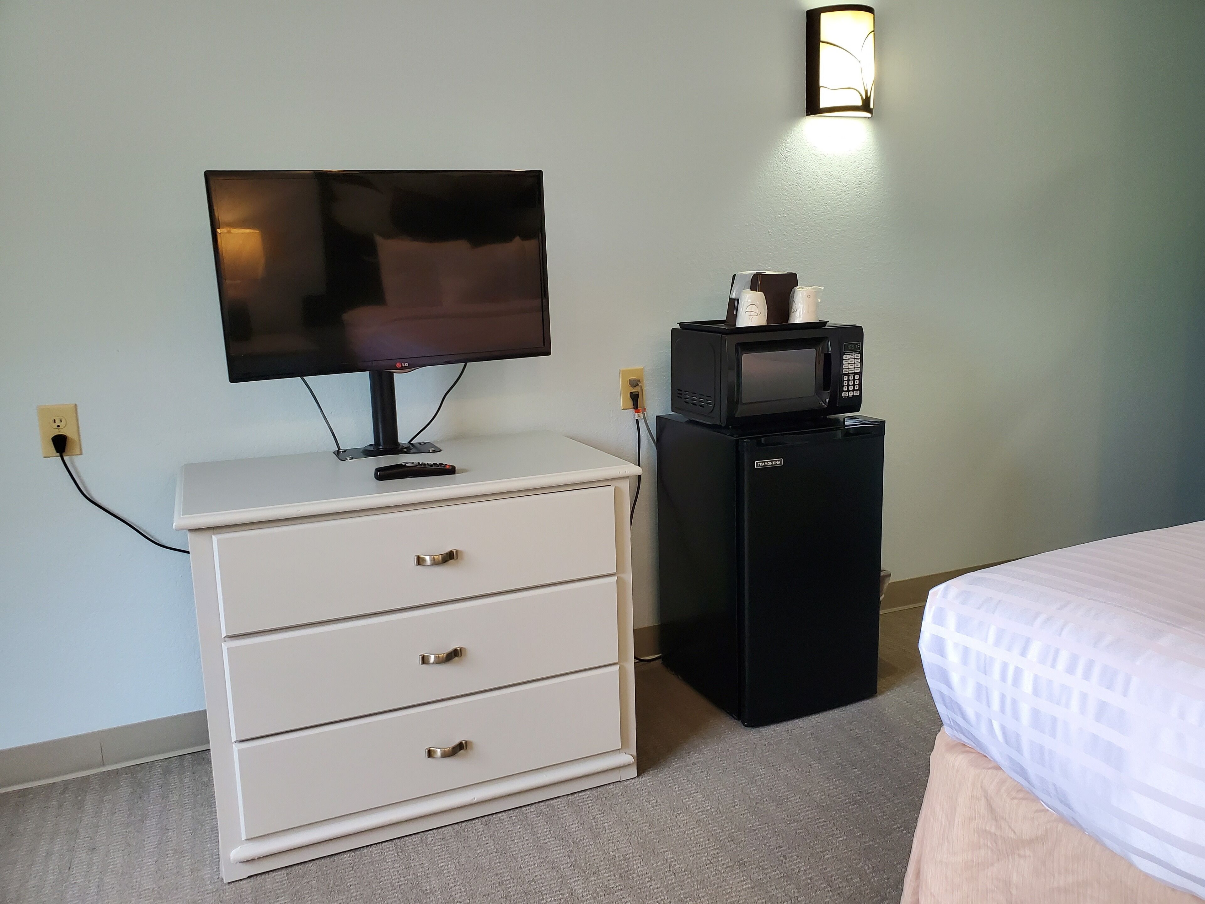 Une chambre d’hôtel avec une commode blanche, un mini-bar noir, une télévision et un lit recouvert d’une courtepointe blanche.