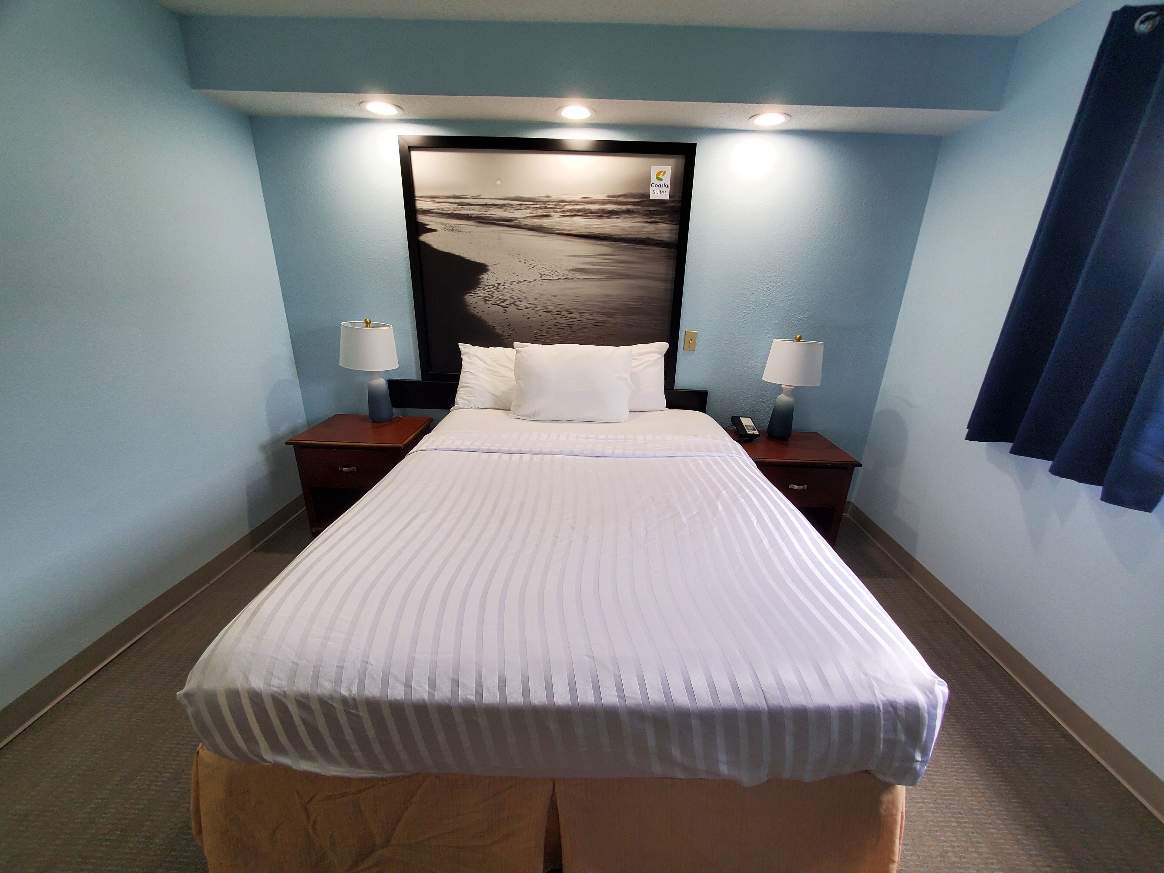 A hotel room with a bed, two bedside tables, two lamps, a nightstand, and a framed picture on the headboard.