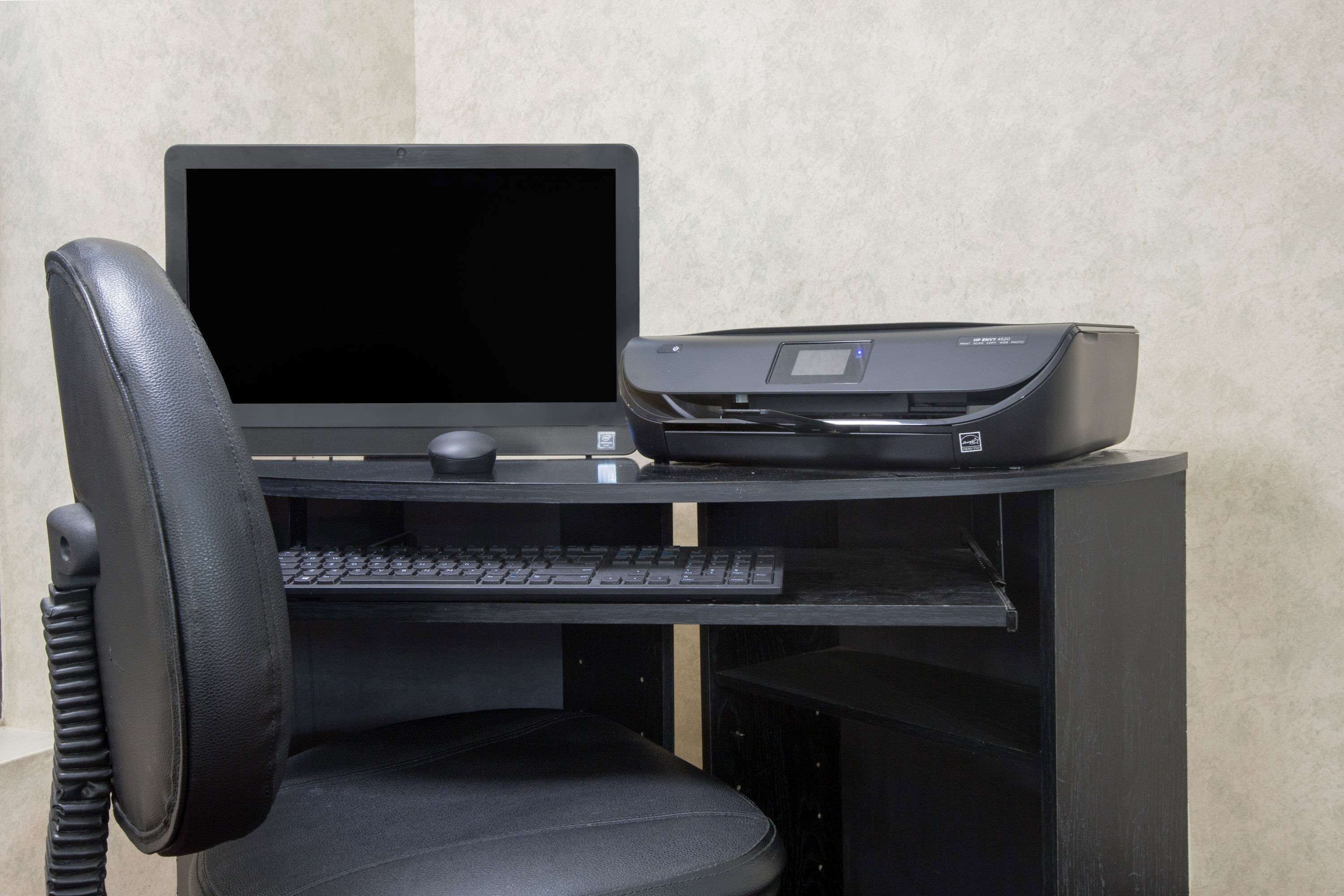 A black office chair with a headrest, a black desk with a flat-screen monitor, a keyboard, a mouse, and a printer.