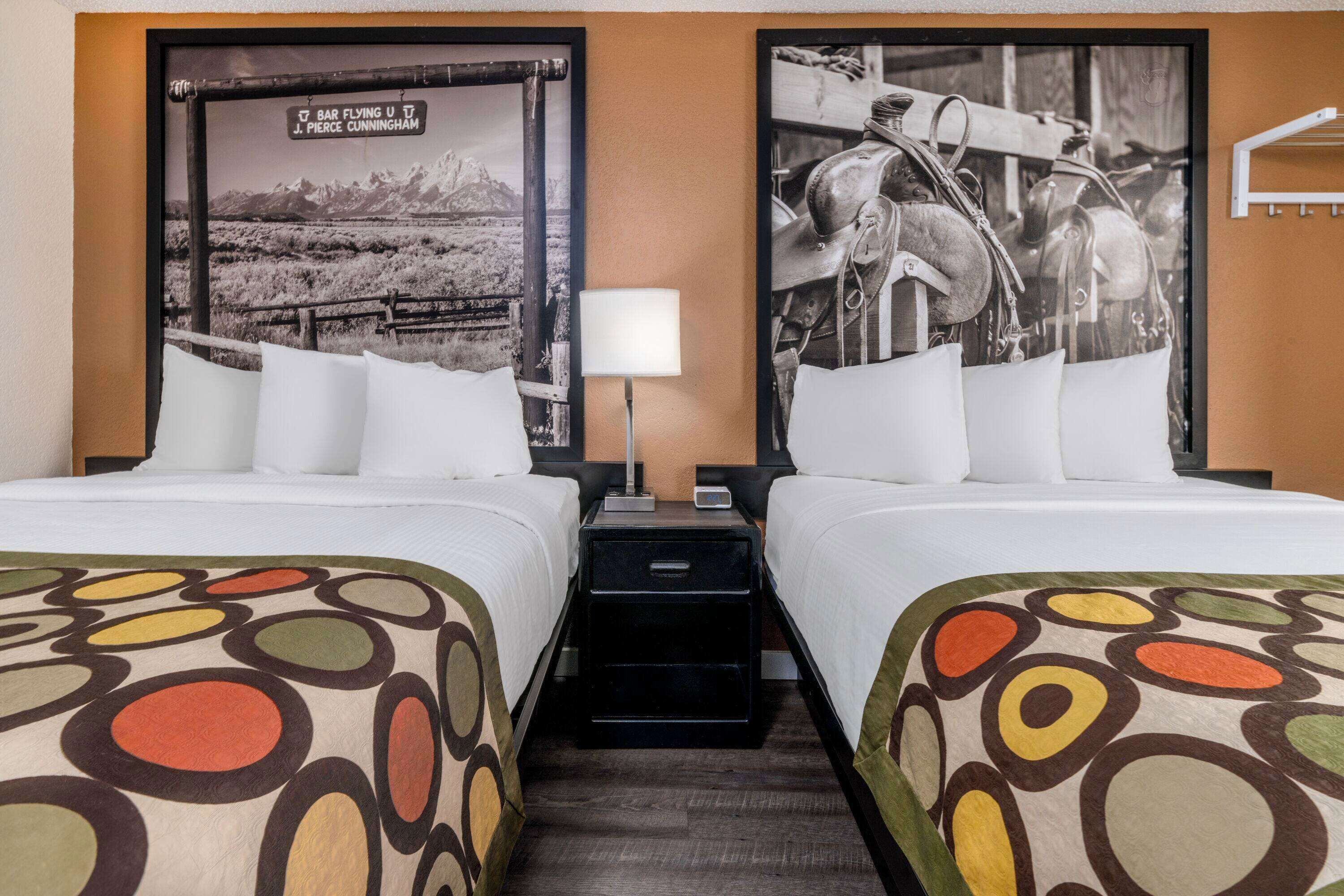 A hotel room with two beds, a nightstand, a lamp, and two posters on the wall.