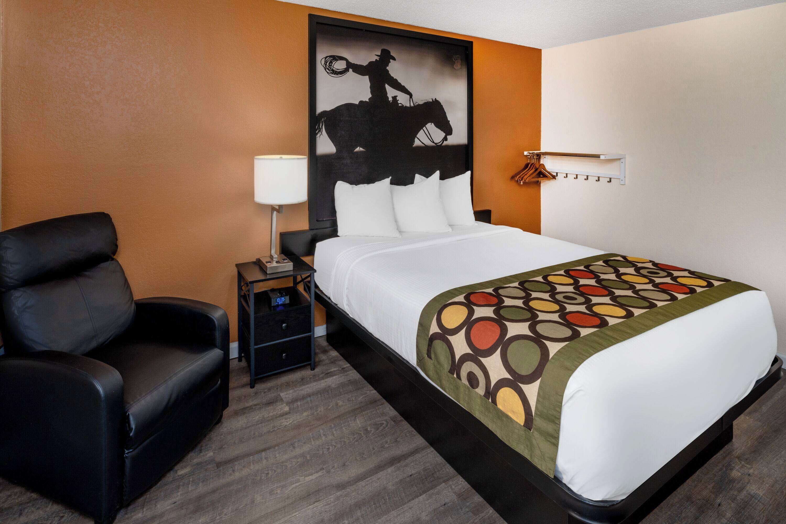 Een hotelkamer met een bed, een zwarte leren fauteuil, een nachtkastje, een lamp en een kapstok aan de muur.