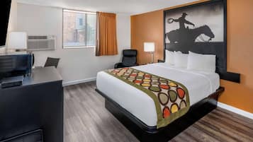 A hotel room with a bed, a desk with a computer, a chair, a window with curtains, and a wall-mounted air conditioner.