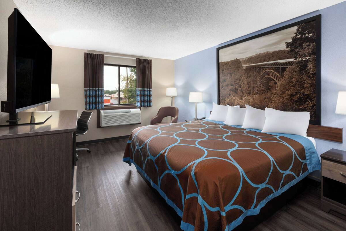 A hotel room with a large bed, a desk with a flat-screen TV, a chair, a window with curtains, and a painting on the wall.