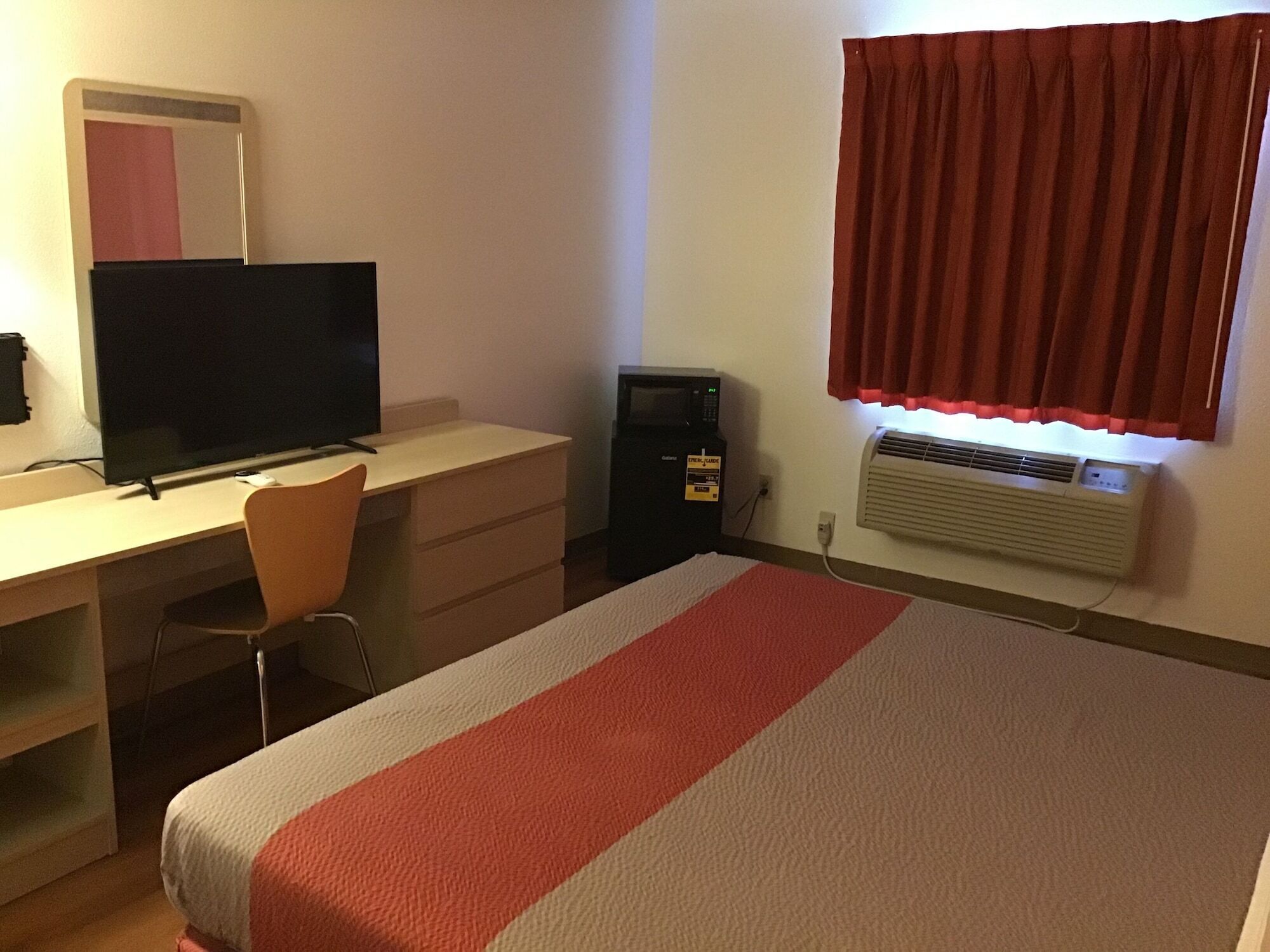 A hotel room with a bed, a desk with a computer, a chair, a TV, a microwave, and an air conditioner.