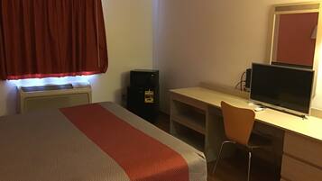 Una habitación de hotel con una cama, un escritorio con computadora y monitor, una silla y un altavoz negro pequeño.